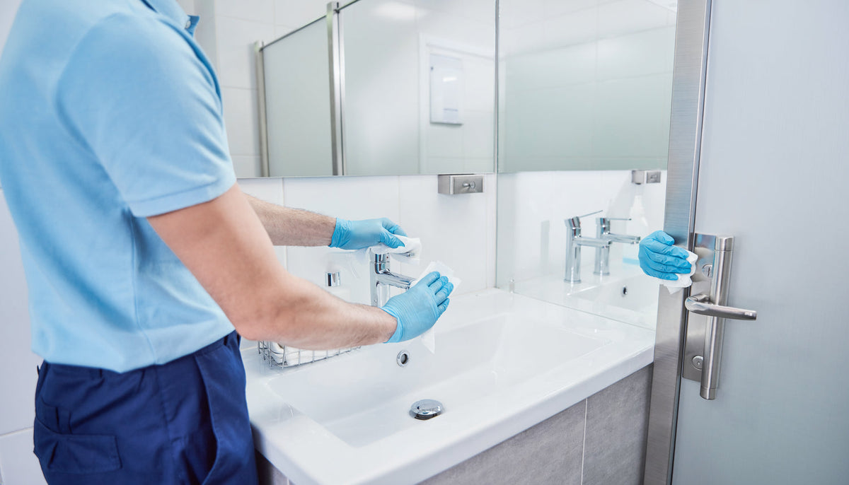 Reinigungskraft reinigt Waschbecken, Armatur und Kontaktflächen in einem öffentlichen Sanitärbereich