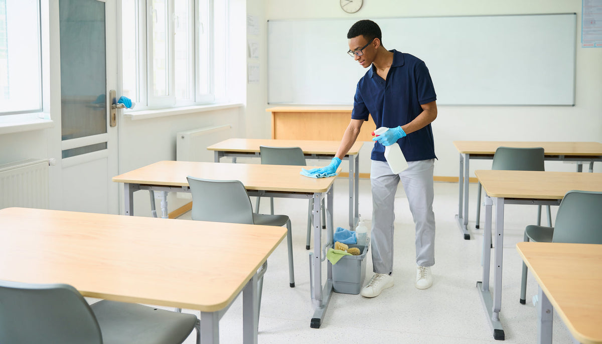 Reinigungskraft reinigt Tische und Kontaktflächen in einem leeren Klassenraum